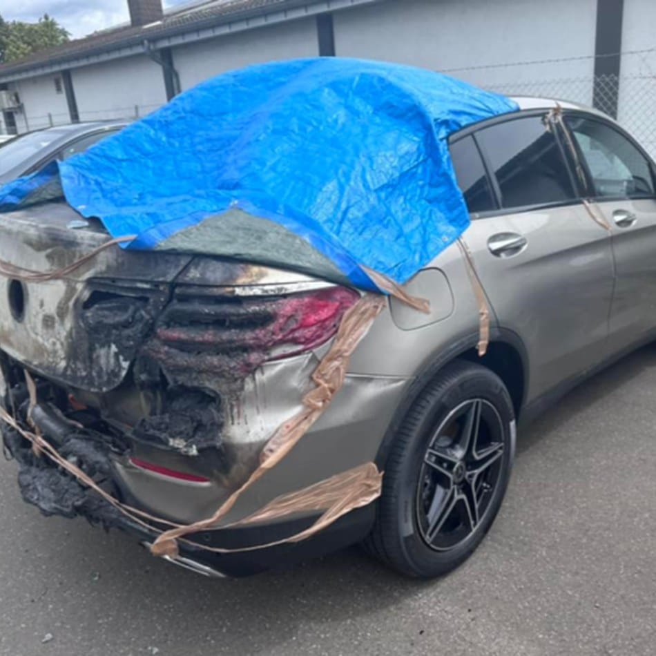 Grauer Mercedes-SUV mit Brandschaden am Heck, Stoßfänger und Rückleuchten zerstört, Fahrzeug mit Plane und Klebeband gesichert